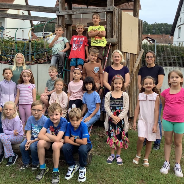 Eine Schulklasse posiert mit zwei Lehrkräften am hölzernen Kletterturm auf dem Schulhof.